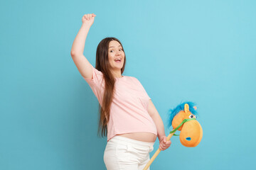 happy young pregnant woman in pink t-shirt on blue background rides  toy horse, family game
