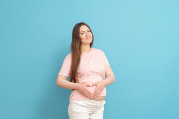 happy young pregnant woman in pink t-shirt on blue background , woman holding heart gesture in her hands