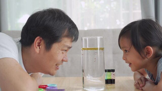 Asian father and little daughter are looking to the color diffusion from oil to water phase experiment, concept of science, education and learning activity for kid. Parent role for homeschooling. 