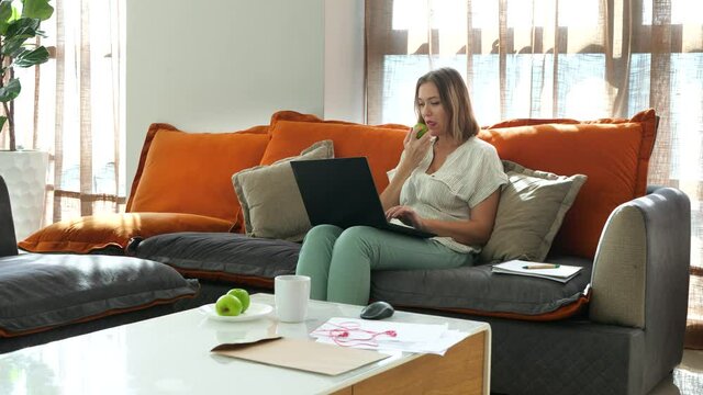 Young woman typing text on laptop and eats apple. Distance job at home room. Orange color pillows on sofa on windows background. Creative professional copywriter. Inspiration time, peace of mind