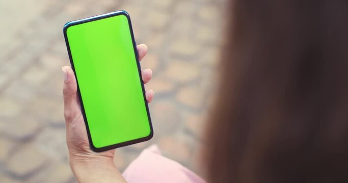 Over shoulder view of female person holding and pressing on smartphone with mockup screen while sitting at street. Concept of chroma key and greenscreen.