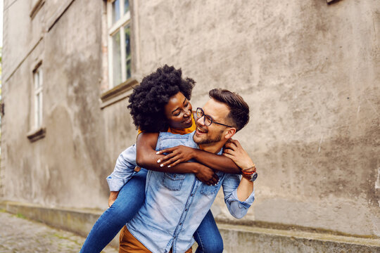 Young smiling overjoyed multicultural hipster couple having piggyback outdoors.