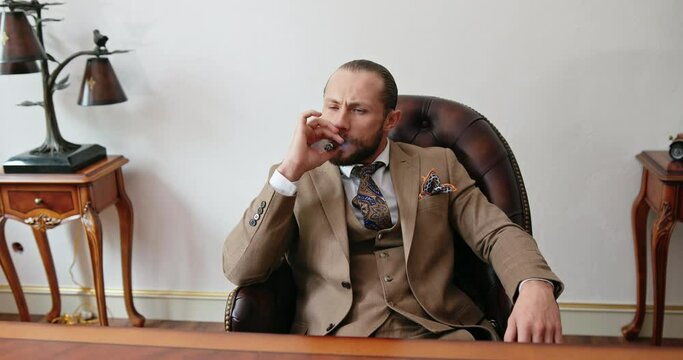 Handsome rich young man in a beautiful expensive suit sits in the office and enjoys smoking a Cuban cigar happily inflating about a successful business