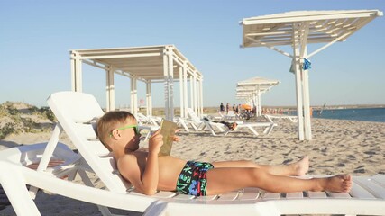 child plays on a tablet on the beach on a Sunny day. A tanned boy in swimming trunks plays on a tablet, resting after a swim. - Powered by Adobe