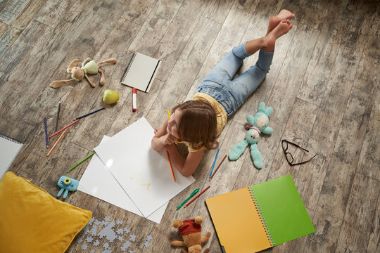 Creative kids concept. Top view of little caucasian girl lying on the wooden floor at home and drawing with colorful pencils on a white sheet of paper