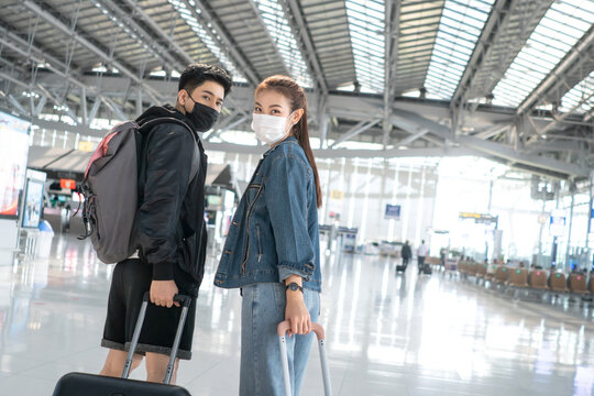 Asian man and woman wearing protective mask in airport terminal for social distancing. new normal friend or couple lifestyle with backpack and luggage ready to travel. Coronavirus prevention