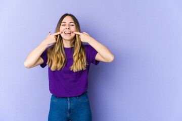 Fototapeta na wymiar Young caucasian woman isolated on purple background smiles, pointing fingers at mouth.