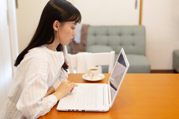 リモートワーク。自宅のキッチンテーブルでノートパソコンでの仕事中、コーヒーを飲む若く美しいビジネスウーマン。