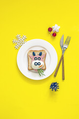 Obraz na płótnie Canvas Funny Christmas sandwich made of bread, sausage, egg and olives in the form of a bull, an edible symbol of 2021 on a white plate decorated with Christmas toys on a yellow background, top view