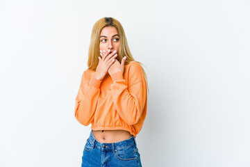 Young blonde woman isolated on white background thoughtful looking to a copy space covering mouth with hand.