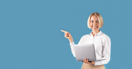 Happy business lady with laptop computer pointing aside at space for your text, blue studio...