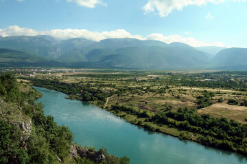 Fototapeta na wymiar Neretva river in a valley near the city of Mostar (Bosnia and Herzegovina) with mountains and a few clouds in the background, being reflected on the water. Neretva is a major river of Balkans