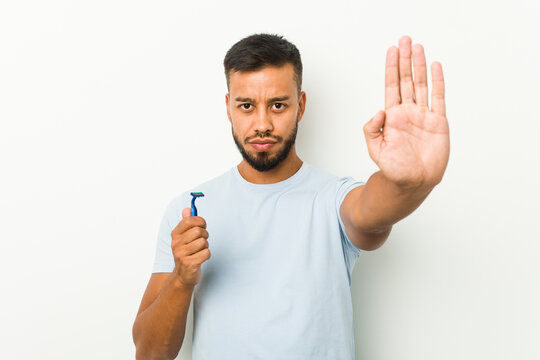 Young south-asian man holding a razor blade standing with outstretched hand showing stop sign, preventing you.