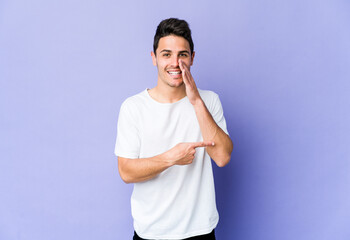 Young caucasian man isolated on purple background saying a gossip, pointing to side reporting something.