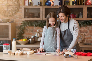 Happy young dad and little daughter baking pie in kitchen, checking recipe