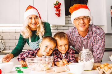 Family of two twins boys and age parents preparing cookies for Chtistmas in light kitchen wearing Santa hats and looking at camera.