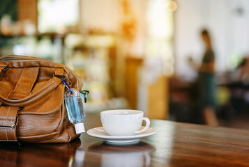 Mini alcohol gel bottle to kill Corona Virus(Covid-19) hang on a brown leather shoulder bag with hot coffee on table in cafe.New normal lifestyle.Health care concept.Selective focus on alcohol gel