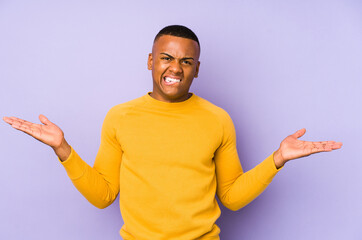 Young latin man isolated on purple background confused and doubtful shrugging shoulders to hold a copy space.