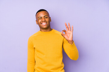 Young latin man isolated on purple background winks an eye and holds an okay gesture with hand.