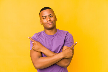 Young latin man isolated on yellow background points sideways, is trying to choose between two options.