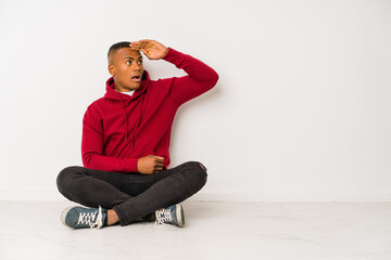 Young latin man sitting on the floor isolated looking far away keeping hand on forehead.