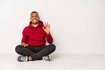 Young latin man sitting on the floor isolated cheerful and confident showing ok gesture.