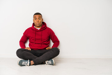 Young latin man sitting on the floor isolated shrugs shoulders and open eyes confused.