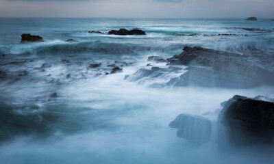 Obraz premium the power our oceans, closeup photography of the waves hitting into the rocks, and the water splashing up into the air during the sunset with beautiful cloudscape in the background 