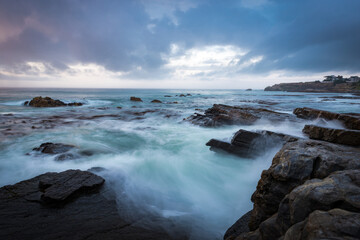 Obraz premium the power our oceans, closeup photography of the waves hitting into the rocks, and the water splashing up into the air during the sunset with beautiful cloudscape in the background 
