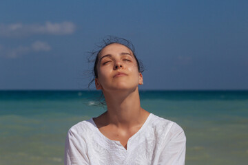 Side view of woman sitting on sandy beach against blue sky outdoors.Portrait of young beautiful girl at the beach.Young woman sitting on a beach and looking at waves.
