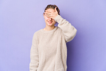 Young caucasian woman on purple background covers eyes with hands, smiles broadly waiting for a surprise.