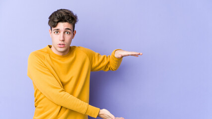 Young caucasian man isolated on purple background shocked and amazed holding a copy space between hands.