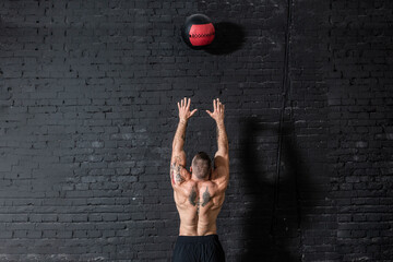 Young strong sweaty focused fit muscular man with big muscles doing throwing medicine ball up on the wall for cross training hard core workout in the gym real people
