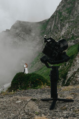 stabilizer with a camera standing in the mountains in cloudy weather, behind him is the groom and the bride in a wedding dress, wedding photography