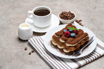portion of Classic tiramisu dessert with raspberries and blueberries, cup of coffee and cream or milk on grey concrete background