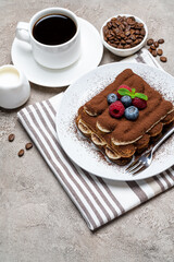 portion of Classic tiramisu dessert with raspberries and blueberries, cup of coffee and cream or milk on grey concrete background