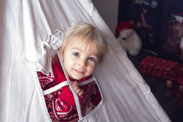 Cute little blonde boy with Christmas sweater, looking from a window of teepee at home