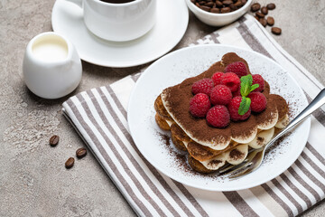 portion of Classic tiramisu dessert with raspberries and cup of coffee and cream or milk on grey concrete background