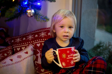 Sweet toddler boy in christmas pajama, sitting on window close to christmas tree, drinking milk