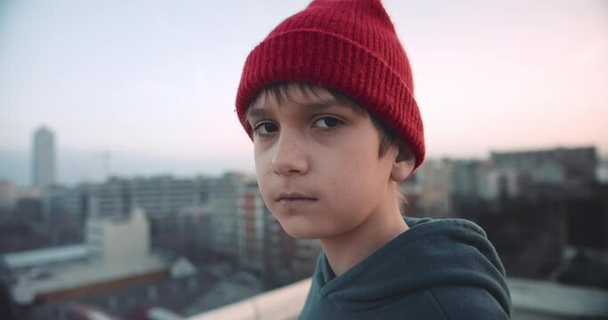 Portrait handsome young boy on rooftop at sunset wearing hoody and beanie red hat looking confident in urban city background.