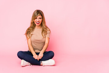 Young blonde caucasian woman sitting on a pink studio screaming very angry and aggressive.