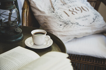 A cup of coffee stands on a vintage table next to a book on the table and an armchair with a pillow. Vintage style. Good morning.