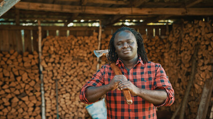 African man lumberjack with the axe standing in front of stack of cutted timber for winter. High quality photo