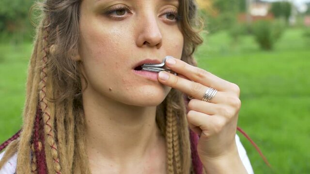 Close-up outdoors portrait of young female shaman with long dreadlocks playing on traditional hutsul drymba, jaw's harp, silver vargan