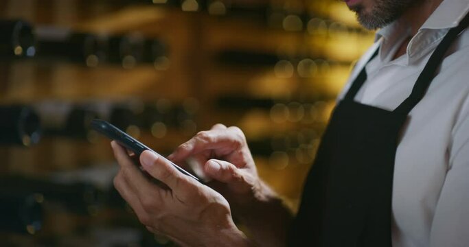 Modern farmer or winemaker is using winery online commerce applications on a smartphone for checking customer service and selling orders summary of his wine production in a wine cellar.
