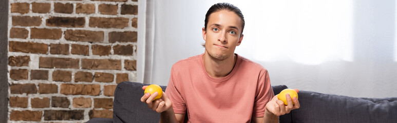 horizontal image of young skeptical man holding lemons while looking at camera at home