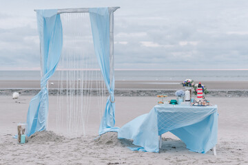 Maritime elegant pastel blue wedding arch and decor table on the beach background. Table with flowers, cackes, candles, mushrooms. Outdoor ceremony decoration concept. Northsea, Germany.