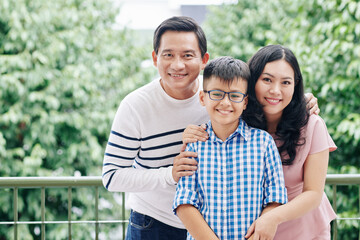 Portrait of joyful Asian mother, father and their preteen kid smiling and looking at camera when standing on apartmernt balcony