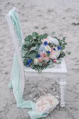 Wedding decoration on the beach, boho white chair with pastell green covers, beautiful big bride bouquet, pink, blue, yellow roses and greenery in sand background. Sea ceremony.
