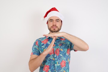 Young caucasian man wearing hawaiian shirt and Santa hat over isolated white background being upset showing a timeout gesture.